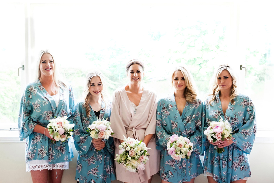 Bridal party smile in front of window at wedding venue with bouquets in hand. All the bridesmaids are wearing blue floral dressing gowns, and the bride wears a blush pink dressing gown. Their makeup is done by Jennifer Louise Studio.